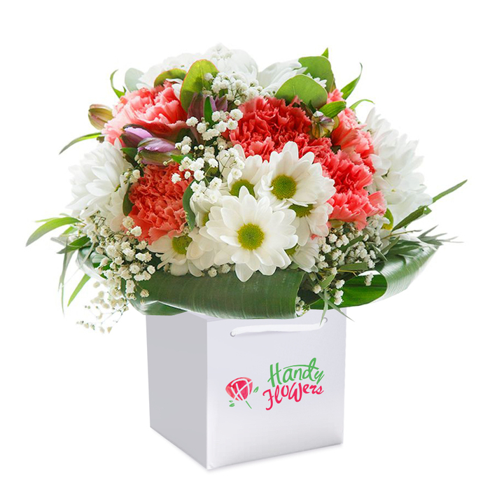 Set against a snowy white background, this vibrant bouquet is rendered in a pristine square gift bag, embodying the freshness of a New Cross morning. Peach and coral carnations dominate the arrangement, their ruffled, multi-layered petals spilling over in gentle waves of colour and texture. White daisies, notable for their bright lime-green centers, nestle among the carnations, shimmering with simple cheerful beauty. The bouquet sparkles with fine sprigs of airy white gypsophila, introducing a dreamy lightness that dances throughout. Tucked behind and to the left, subtle buds edged in purple-pink tones add a layer of mystery and depth, hinting at more blooms soon to come. Broad, deep green leaves wrap the base, fanning out in glossy freshness and completing the composition with a reassuring solidity. The white bag is topped with thoughtfully chosen ribbon handles, ready for easy carrying to addresses across New Cross, from Avonley Road to the creative corners near Deptford. This image not only speaks to the attention and care given by local florists, but also to the special moment when a bouquet is given-a spark of colour and life shared across the community. Perfect for birthdays, anniversaries, or as a thoughtful gesture, this arrangement showcases the artistry of New Cross floristry in every detail.